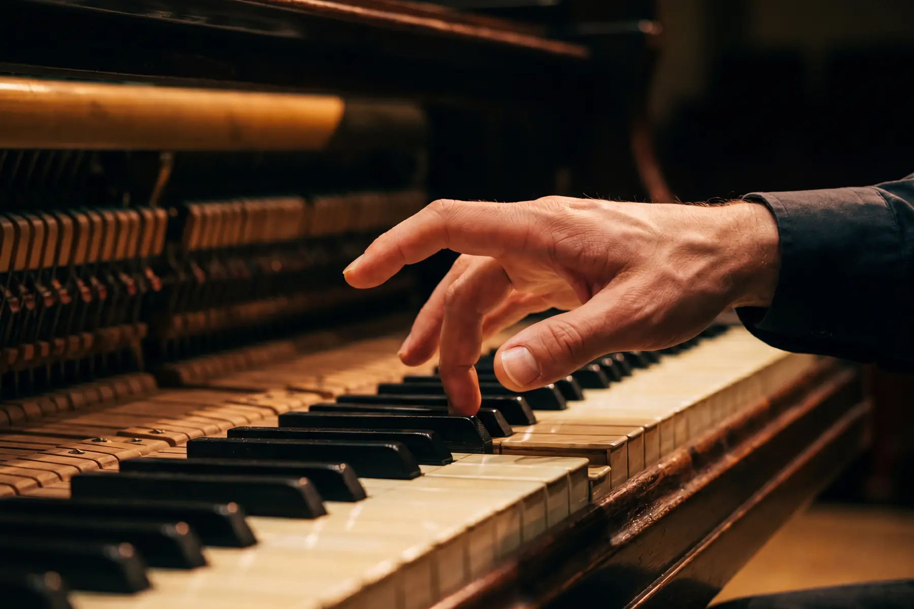 A single hand above a vintage upright piano keyboard with the wrist and fingers in a high curved arch — one finger contacting a black key while the others remain elevated in the preparatory learning position