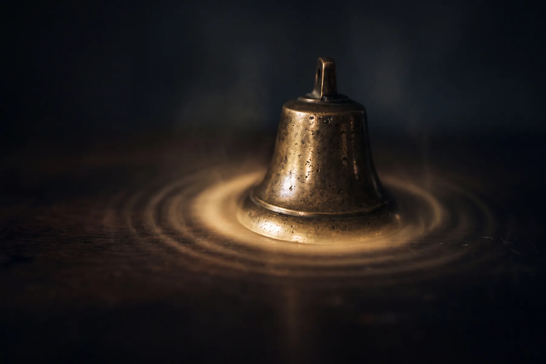 A small antique brass bell, just struck, with a warm golden halo and faint rings of light radiating outward from its rim into the darkness, thinning as they expand — resonance caught the instant it begins to fade.
