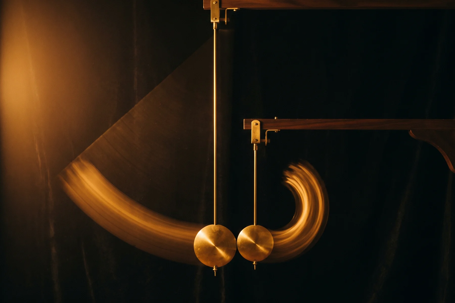 Two brass pendulums of different lengths, one hung high and one hung low, arrive side by side at the bottom of their swing at the same instant, a wide slow golden arc trailing one and a tight quick arc trailing the other against deep darkness.