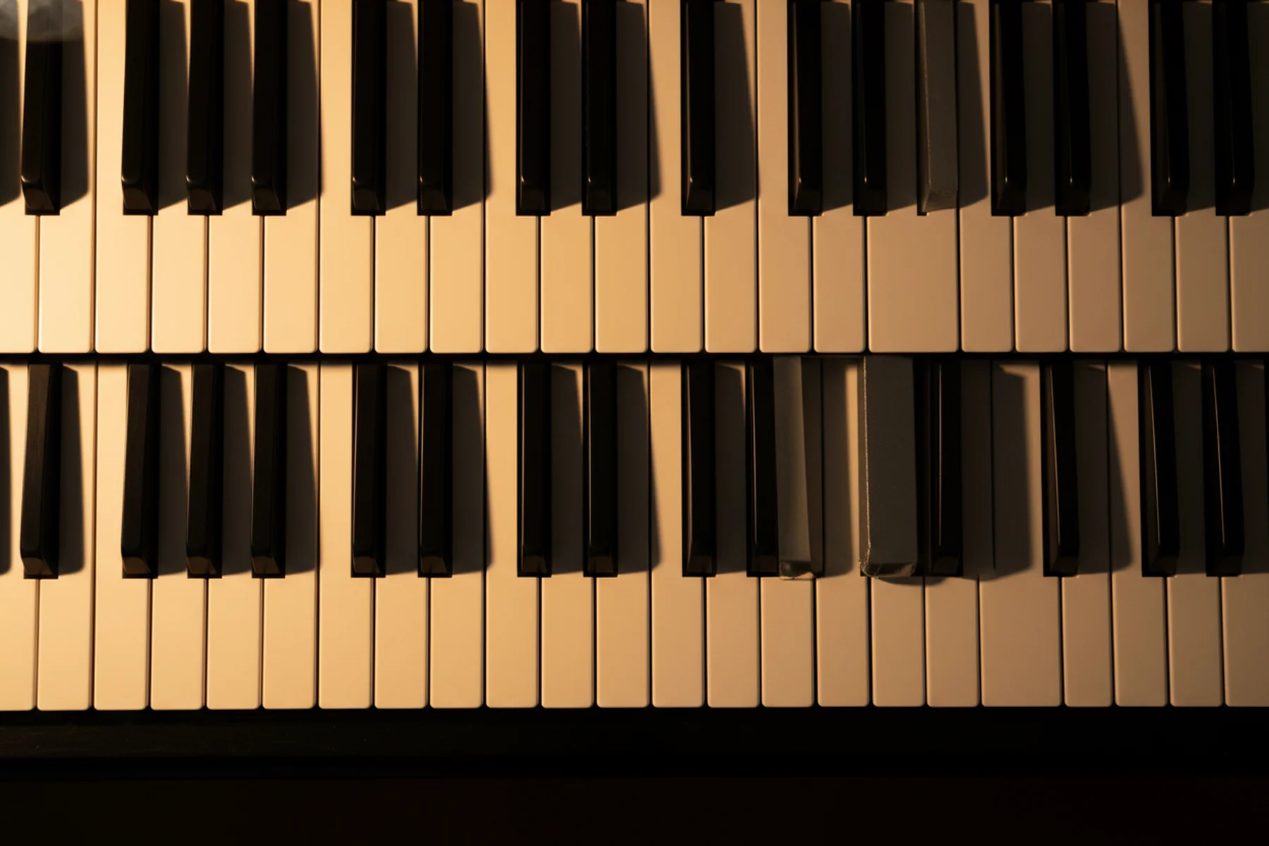 Close overhead view of piano keys bathed in warm amber light, the rows of white and black keys filling the frame in clean repeating geometry — the even terrain a scale must navigate