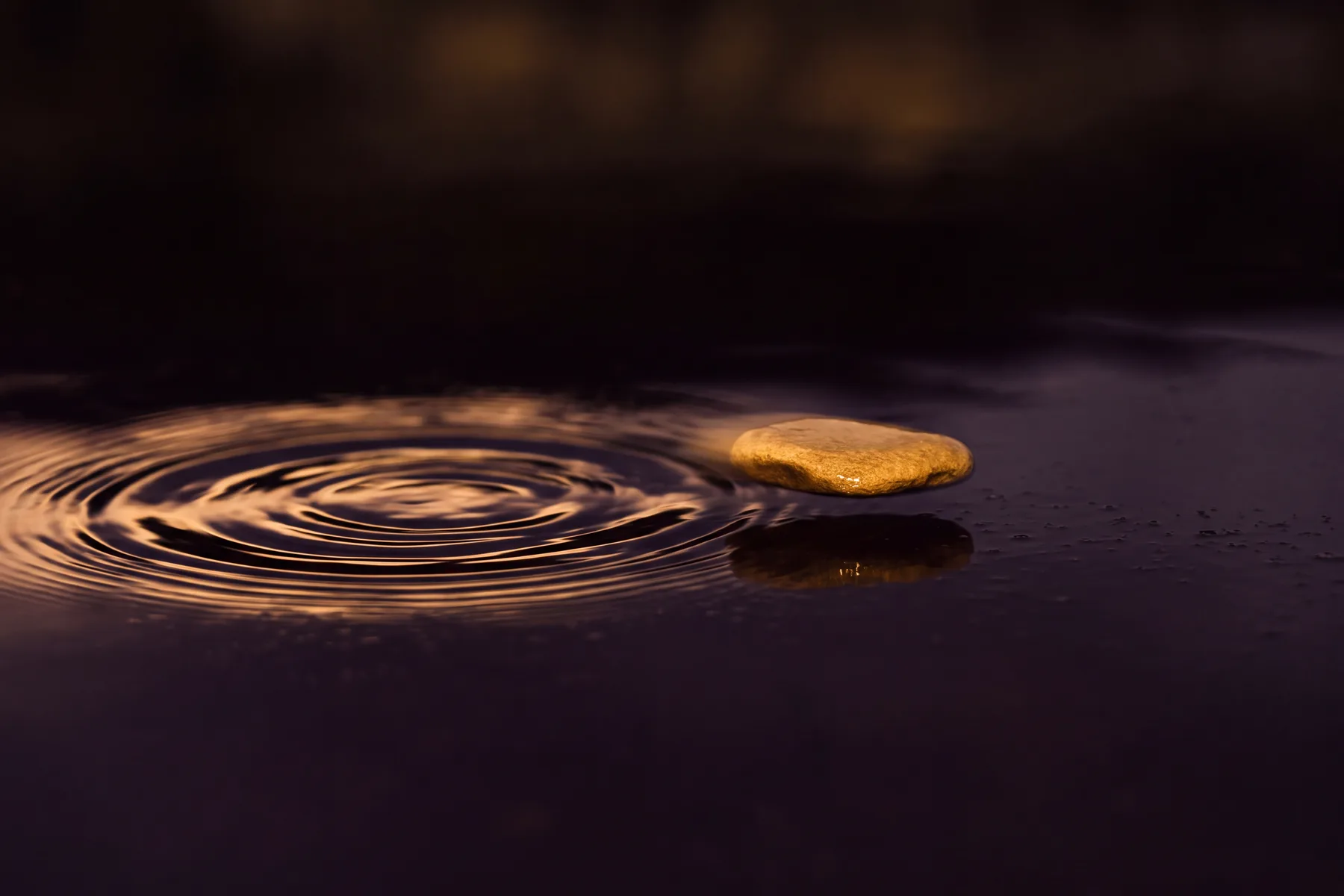 A golden-lit stone mid-flight, already skipping toward its next point of contact on dark water, while the ripple from its previous touch is still visibly expanding behind it.