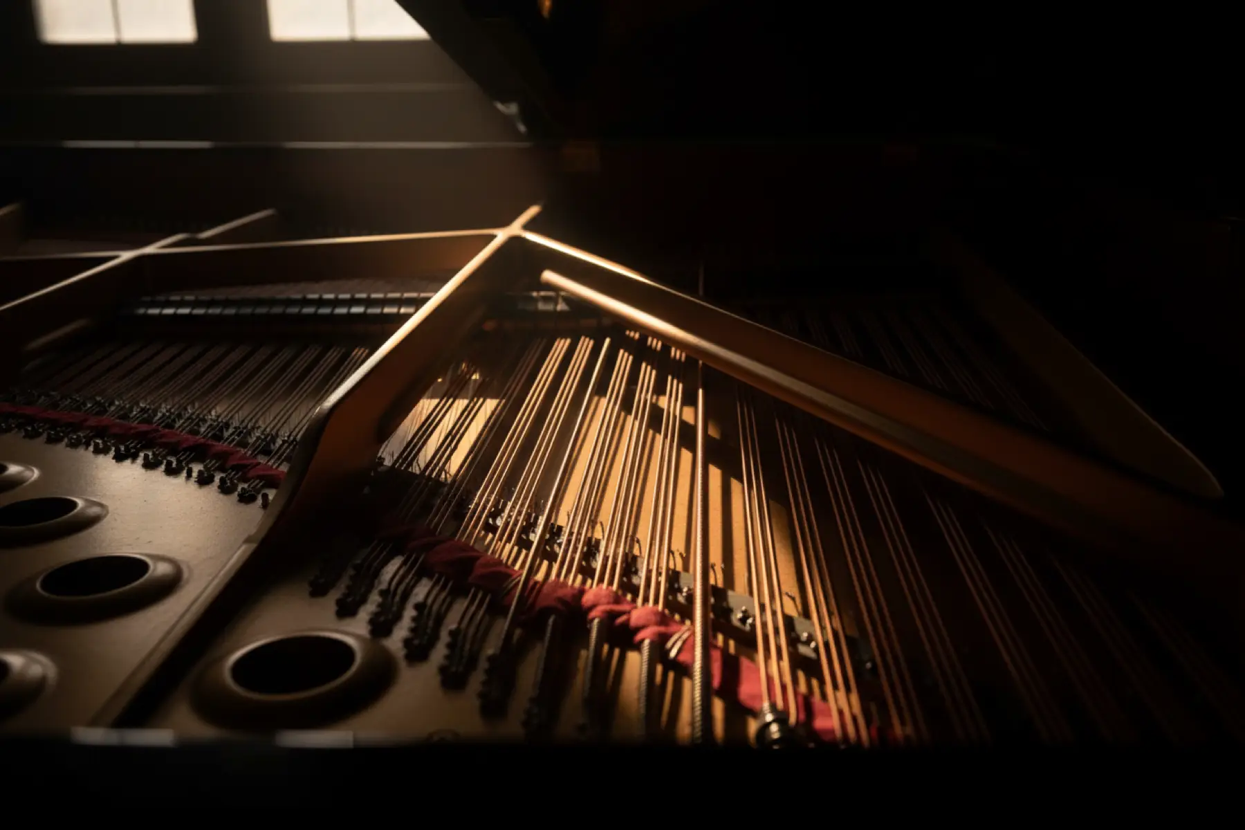 Grand piano interior in low-key light, a diagonal shaft of amber illuminating cross-strung bass strings — the interconnected mechanism of arm, wrist, and finger coordination