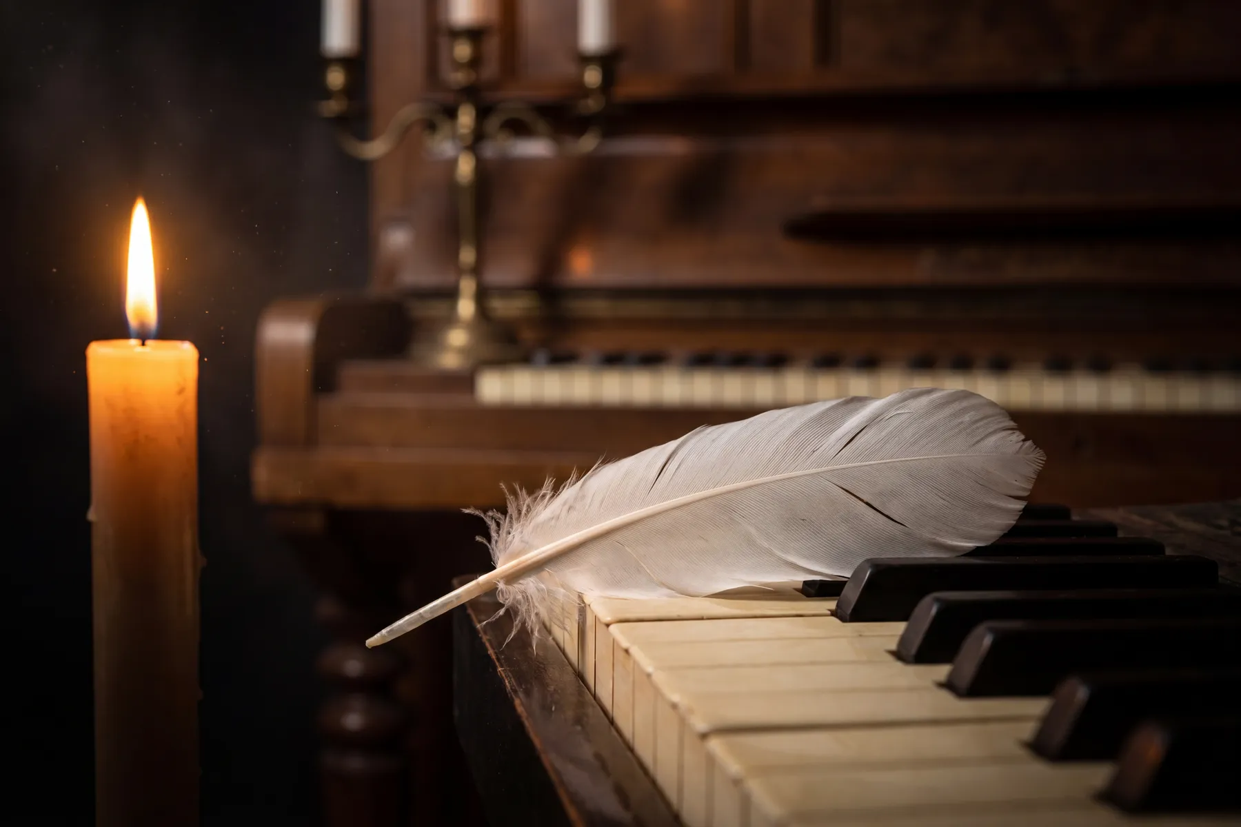 A white feather resting across the ivory and ebony keys of an antique upright piano in warm candlelight, a lit candle on the left and ornate brass candlestick holders visible in the background — softness and delicacy at the keyboard
