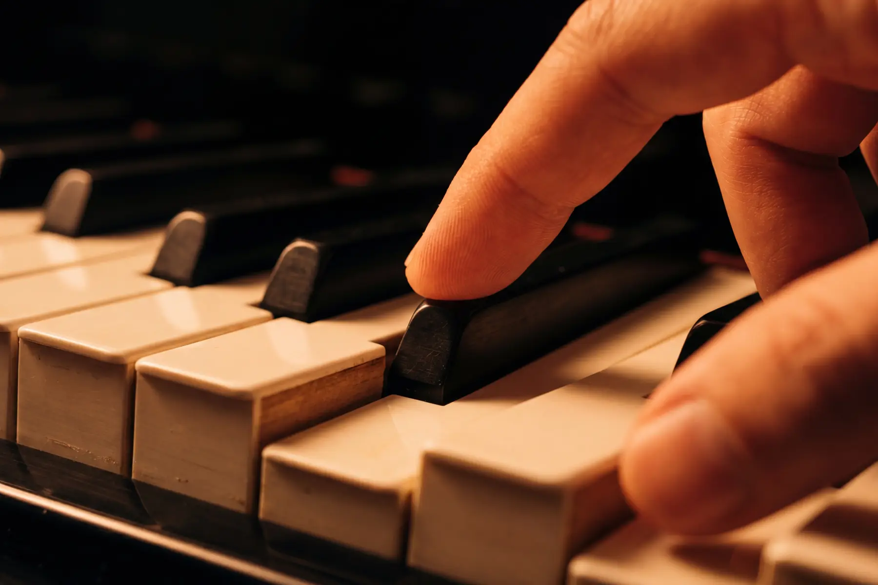 A fingertip arriving precisely onto a black piano key from above — the narrow raised ebony surface flanked by white keys on both sides, the black key's distinct geometry making the targeting precision visible