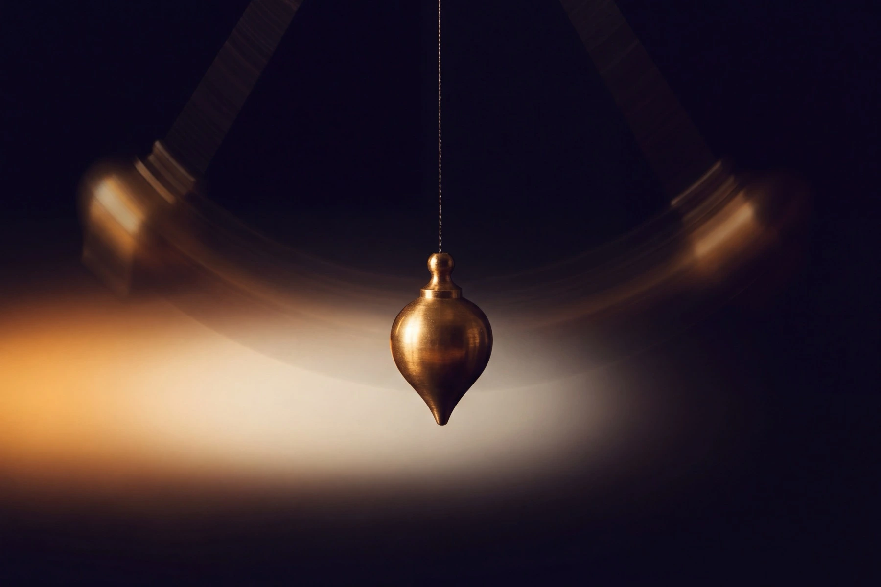 A brass pendulum bob glowing warm gold at the exact bottom of its swing, soft motion-blur arcs tracing its path up and back through deep near-black darkness, evoking momentum gathering toward one low point and easing away again.