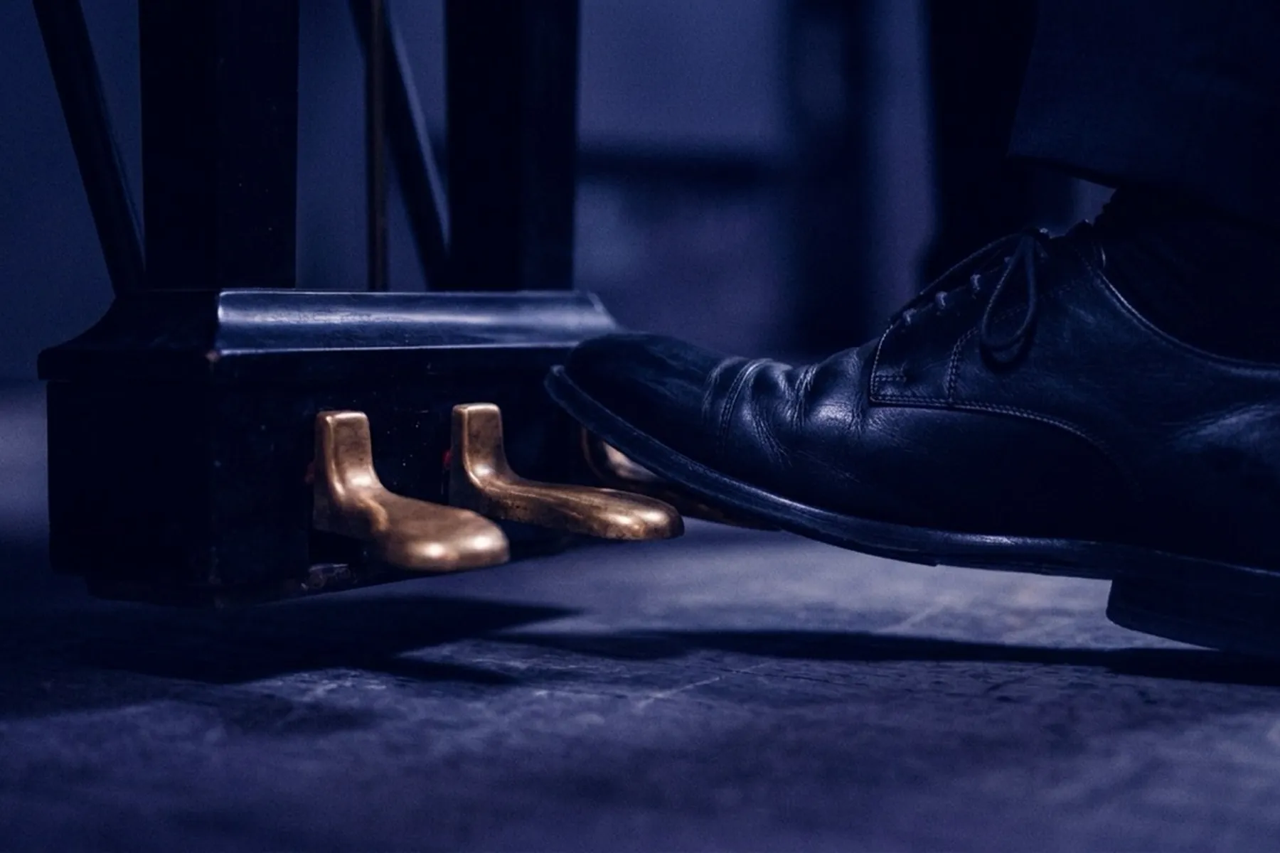 A polished black dress shoe resting on the gold sustain pedal of a grand piano in blue-toned low light — the foot positioned to listen and respond rather than count