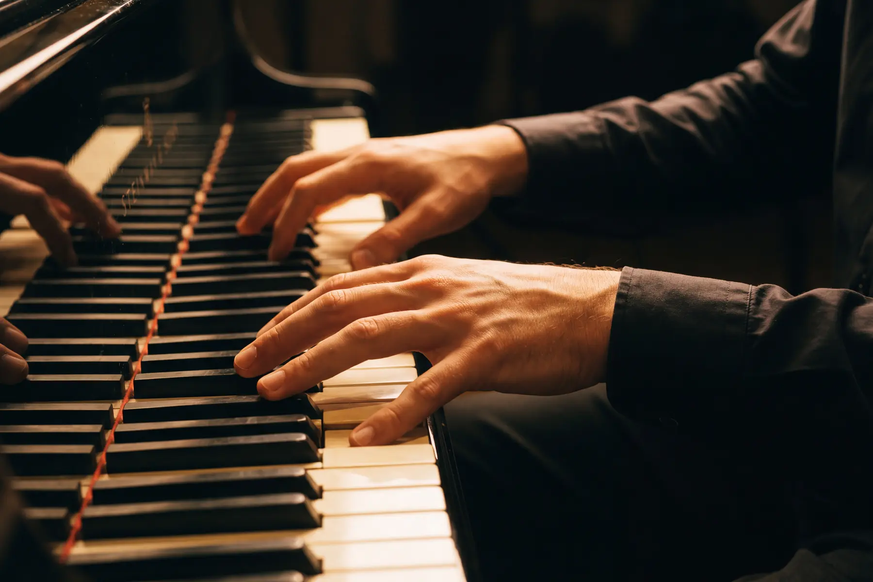 Two hands placed on the keys of a grand piano in warm amber light, arms approaching the keyboard from a natural angle with the wrists at their natural resting level